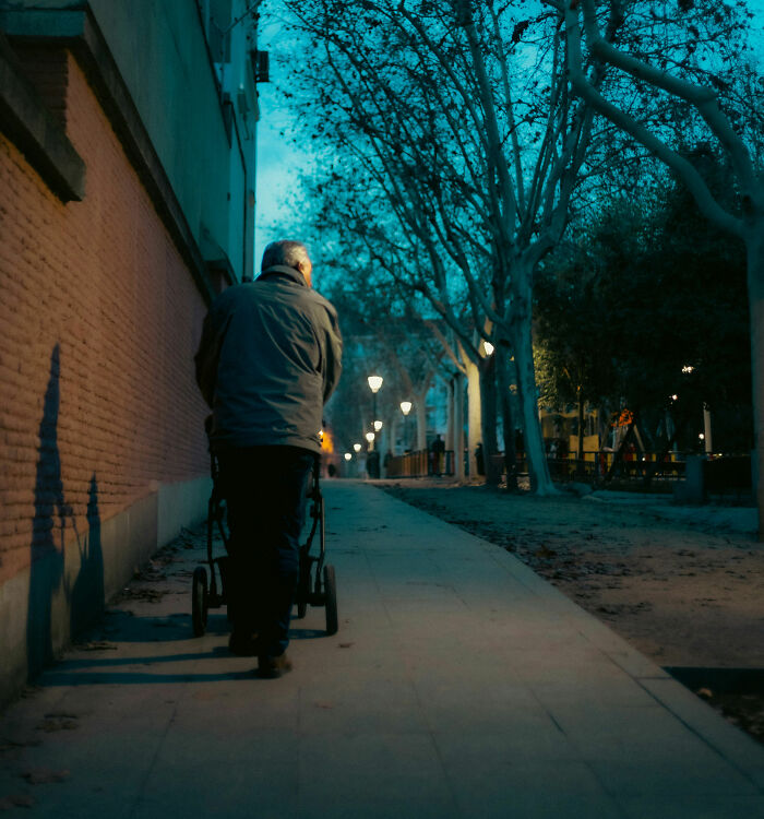 Elderly man pushing stroller along dimly lit urban sidewalk lined with trees during early evening city walk.