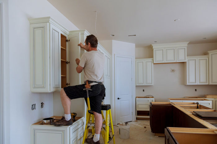 Man working on white kitchen cabinets, adjusting doors to reduce noise from slamming, using a ladder and tools.