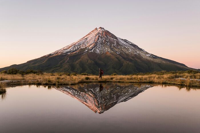 A lone hiker walks along a lake with a snow-capped mountain reflecting in the water, offering a gentle reset.