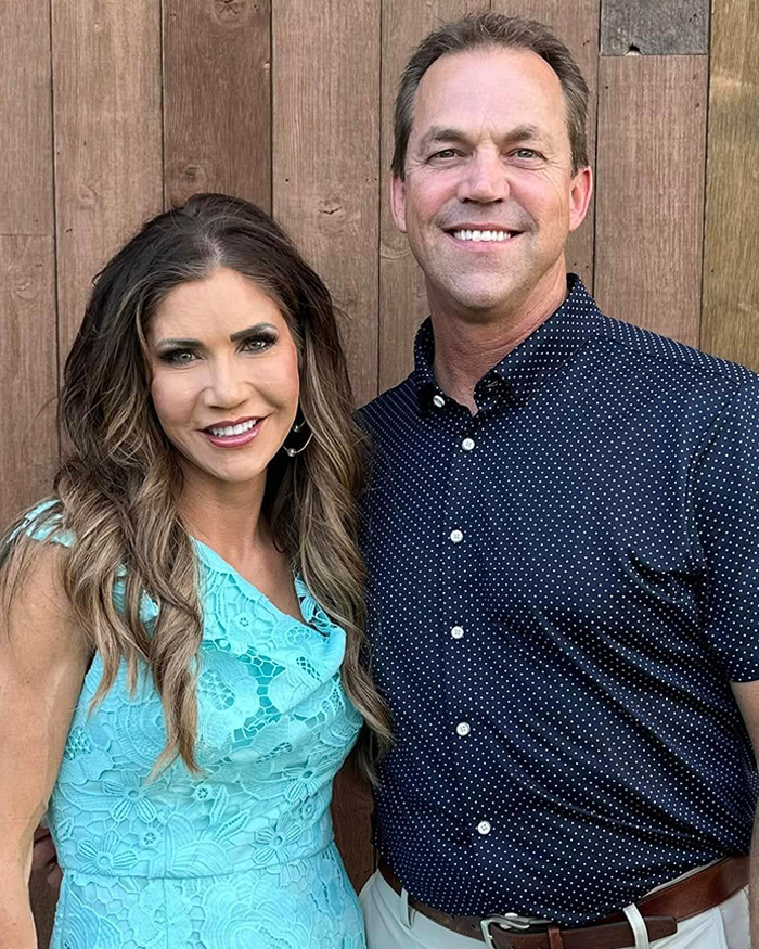 Couple posing in front of wooden wall, linked to Kristi and Bryon Noem $2.6 million debt scandal and luxury spending concerns.