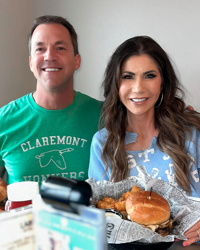 Kristi and Bryon Noem smiling at a restaurant with a burger and tater tots, linked to luxury spending scandal.