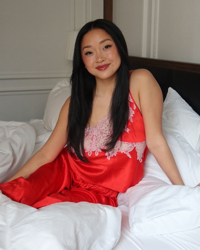 Lana Condor sitting on a bed wearing a red lace satin outfit, smiling with long black hair in a bright room. Lana Condor sitting on a bed wearing a red lace satin outfit, smiling with long black hair in a bright room.
