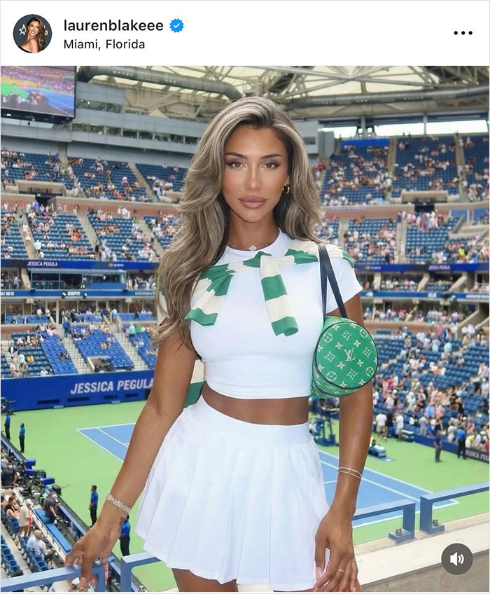 Glamorous white influencer posing at tennis stadium wearing white outfit with a green purse as fans watch the match.