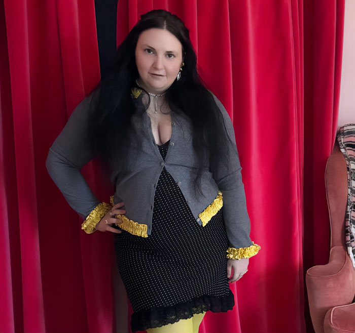Person posing confidently in a black dress and gray cardigan with yellow accents against a red curtain backdrop, symbolizing peak main character syndrome.