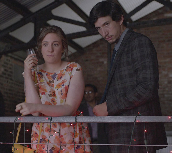 Lena Dunham and Adam Driver on set, portraying characters from the show Girls in a dimly lit scene.