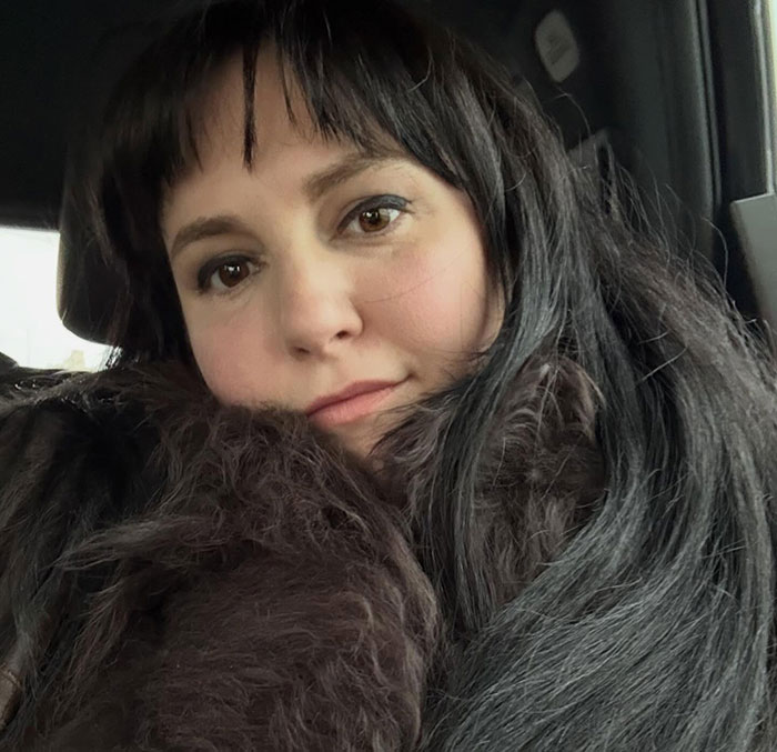 Lena Dunham close-up portrait inside a car, wearing a dark fur coat with natural makeup and long dark hair.