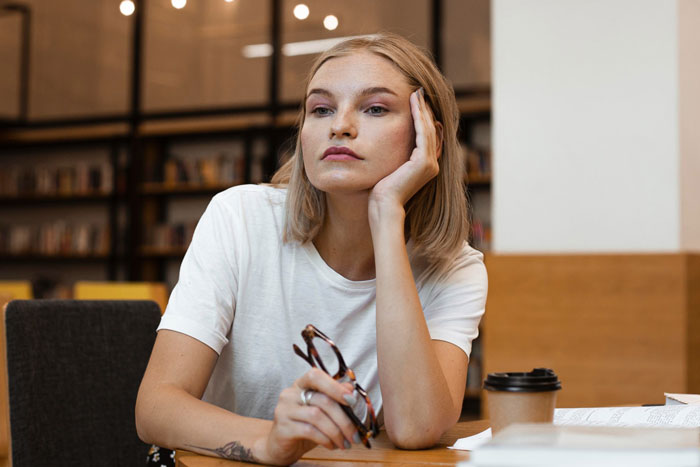 Blonde woman with glasses in hand, resting her chin, deep in thought at a table. Relevant to locking school office security.
