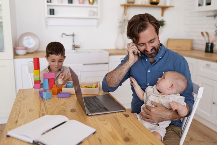 Man holding baby and working at home, while young boy plays with blocks, highlighting family challenges and parenting struggles.