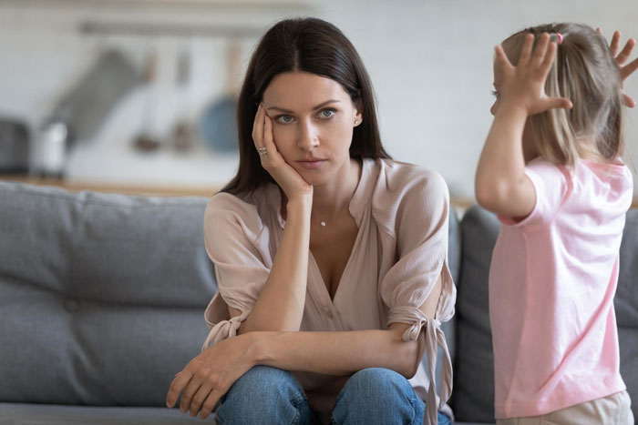 Woman looking stressed sitting on a couch while a child raises hands, reflecting family conflict and abandoned kids at home.