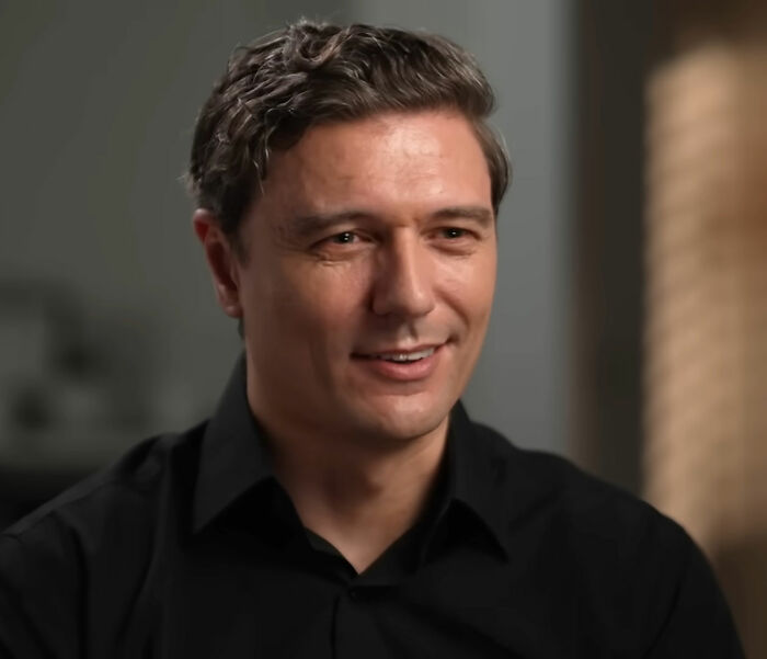 Man with dark hair and black shirt sitting indoors, representing Looksmaxxer in a candid moment during an interview.