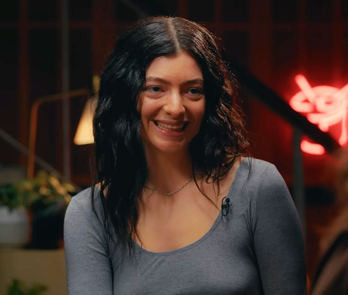Lena Dunham smiling with wavy black hair, wearing a gray top in a warmly lit room discussing main character syndrome.