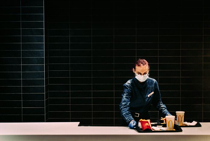 A worker in a mask and blue shirt prepares fast food on trays, evoking anonymous confessions and group therapy.