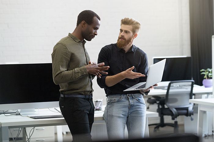 Two men discussing work in an office. One holds a laptop, gesturing. This situation reflects a toxic bestie scenario.