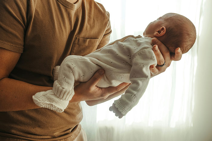 Man in brown shirt gently holding a newborn baby dressed in white near soft natural light background.