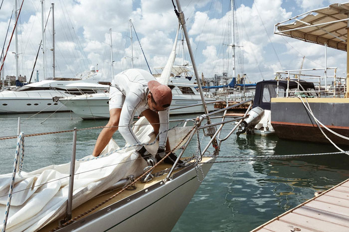 A man in a red cap and white activewear works on the sail of his boat in a sunny marina, destroying his new life.