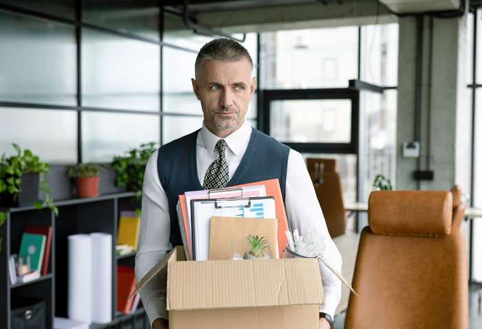 A grey-haired man in a white shirt and vest carries a box of personal items, having his new life destroyed.