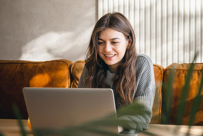 A smiling woman with long brown hair, wearing a gray sweater, looks at her laptop. Is she plotting revenge?