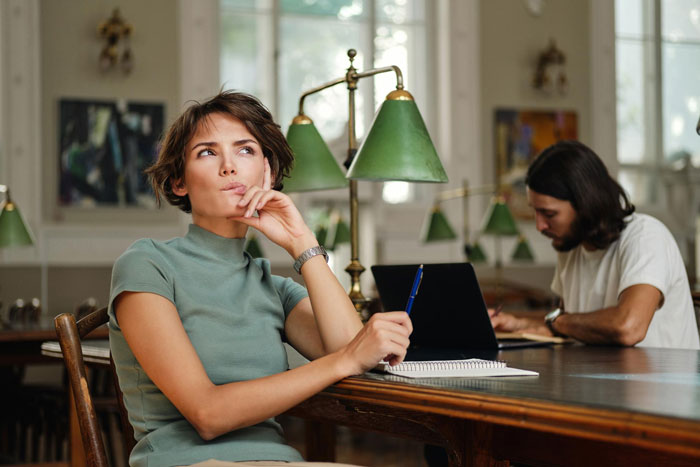 A woman in a green shirt, looking thoughtful while holding a pen. She's destroying his new life.