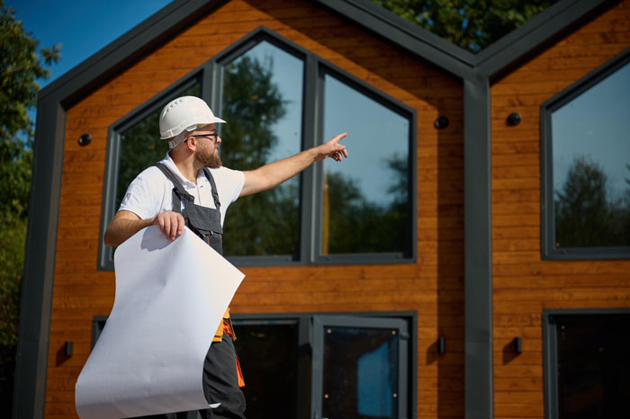 A construction worker in a hard hat and overalls, holding blueprints, points at a modern wooden house, his new life.