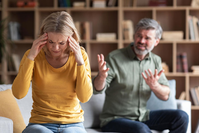 A distressed woman with a headache while a man argues in the background. Illustrates "Guy Dumps Wife To Marry Mistress" revenge story.