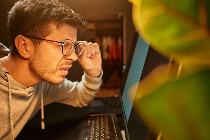 A man with glasses and a grey hoodie squints at a laptop screen, looking surprised, perhaps finding out he was being cheated on.