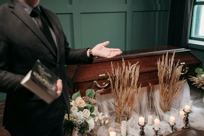 A person in a black suit gestures toward a coffin adorned with flowers and candles, reflecting serious reasons to fire someone.