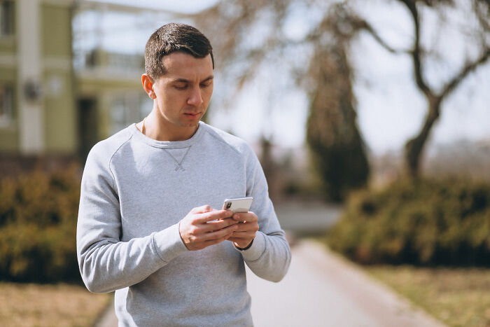 Man in grey sweatshirt focused on his phone outdoors, possibly reading about wildest reasons to fire someone.