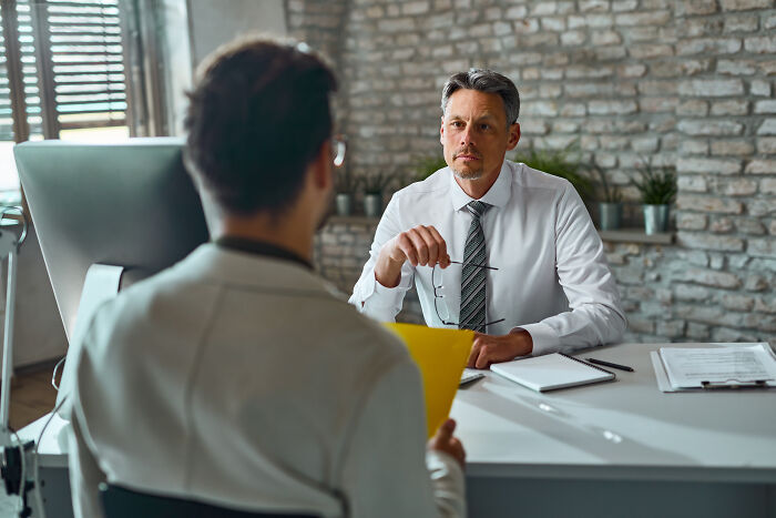 A serious employer in a white shirt and tie, holding glasses, looks intently at an employee, discussing reasons to fire someone.