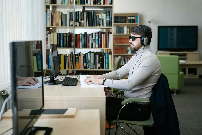Visually impaired man with headphones using Braille device at library desk, highlighting reasons to fire someone.