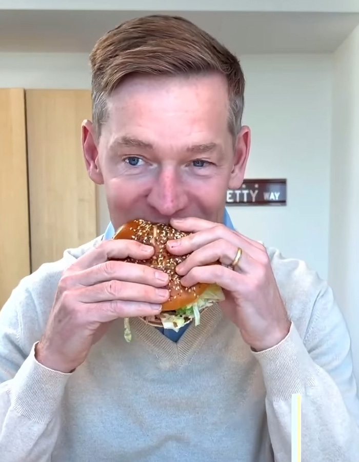McDonald&rsquo;s CEO taking a bite of a Big Arch burger, reacting to the viral sensation in a casual indoor setting.