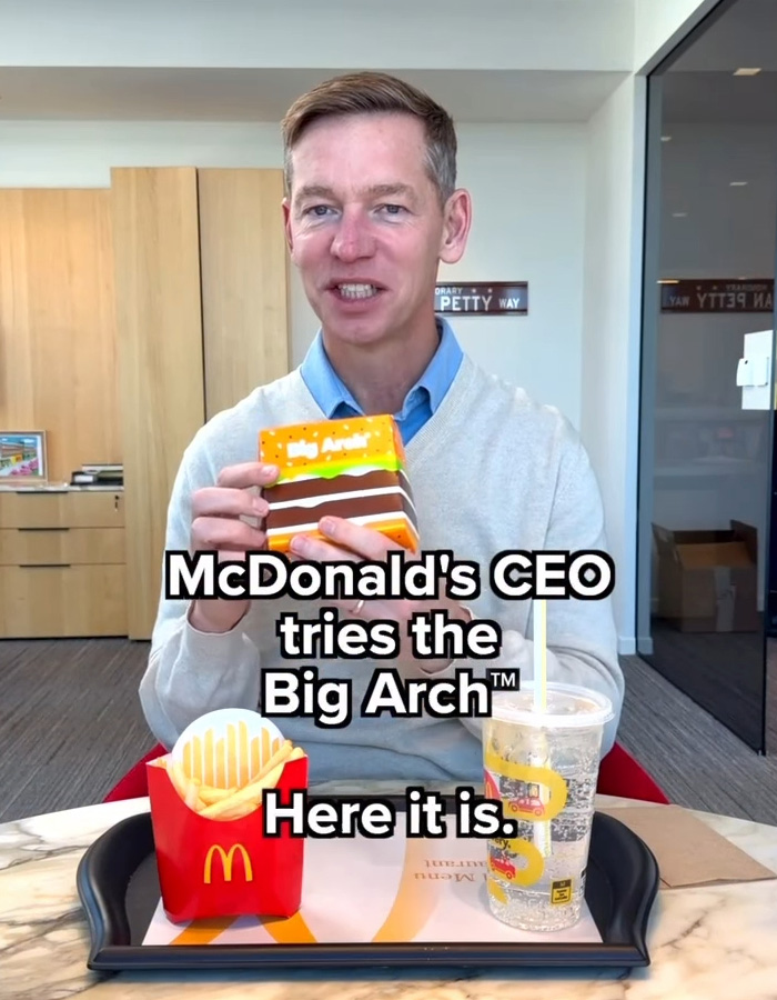 McDonald's CEO holding Big Arch burger box, sitting at a table with fries and a drink in an office setting.