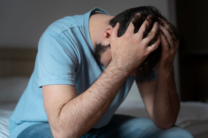 Man in a blue shirt sitting on a bed, holding his head in frustration, reflecting on life consequences of cheating.