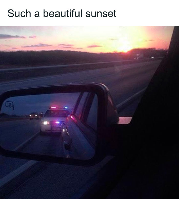 Rearview mirror showing police car with flashing lights behind during a sunset, a funny and lighthearted safe-for-work meme.