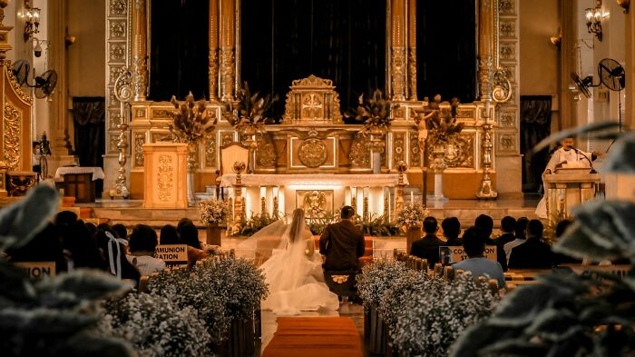 A bride and groom kneeling at the altar during a wedding ceremony witnessed by guests in a decorated church interior.