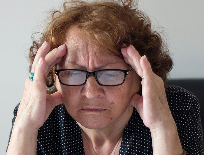 Older woman wearing glasses, looking stressed and frustrated, holding her temples with both hands indoors.