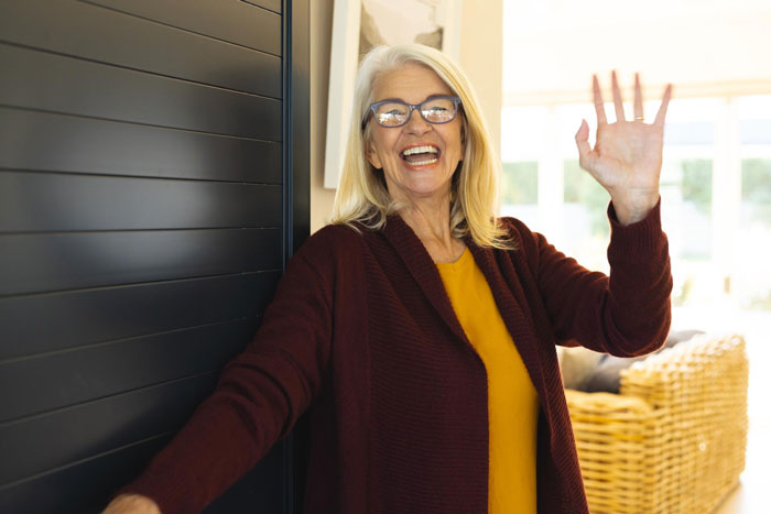 Older woman using emergency key to enter home, smiling and waving, depicting unexpected family drop-in situation.