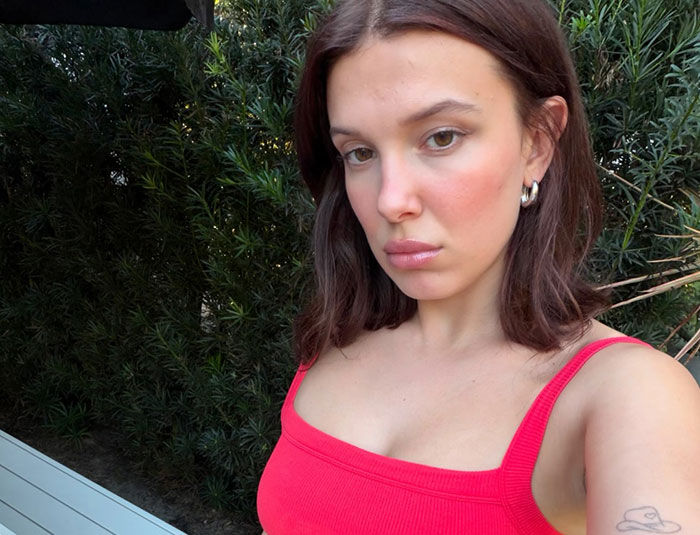 Millie Bobby Brown in a red top against foliage, hinting at Enola Holmes 3 details and a potential major blunder.
