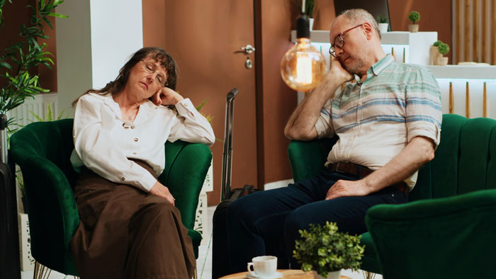 Middle-aged couple sitting apart in green chairs looking upset in a home, illustrating toxic MIL refusing to leave visit.