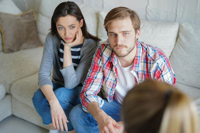 Young couple sitting on a couch looking frustrated during a tense discussion about toxic MIL refusing to leave their home.