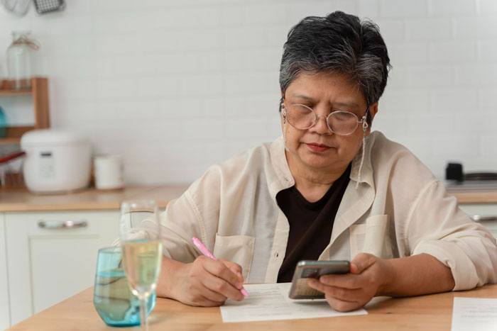 Elderly woman with glasses sitting at kitchen table, writing and looking at phone, representing toxic MIL visiting daughter's home.