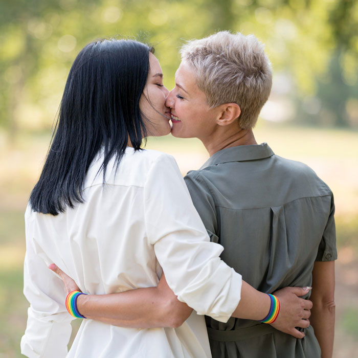 Duas mulheres se abraçando e se beijando ao ar livre, usando pulseiras de arco-íris simbolizando o orgulho e o amor LGBTQ.