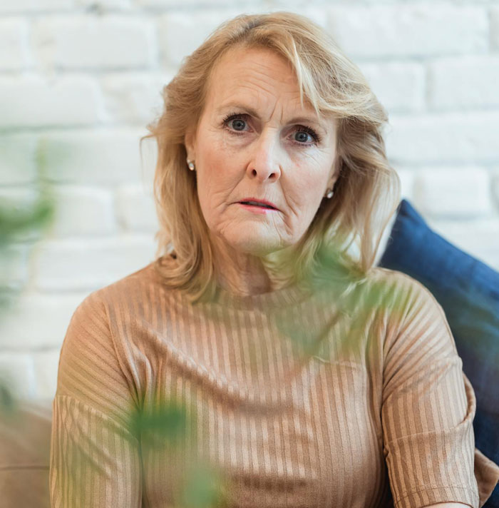 Mulher mais velha com expressão preocupada, sentada dentro de casa, refletindo o dilema sobre a mãe controlar o arranjo de dormir com a futura esposa