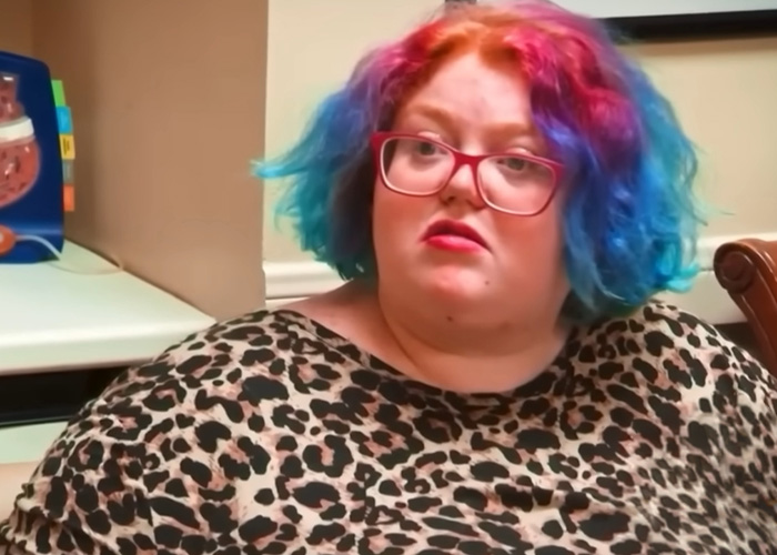 Dolly Martinez from My 600-Lb Life with colorful hair and red glasses wearing a leopard print top indoors.