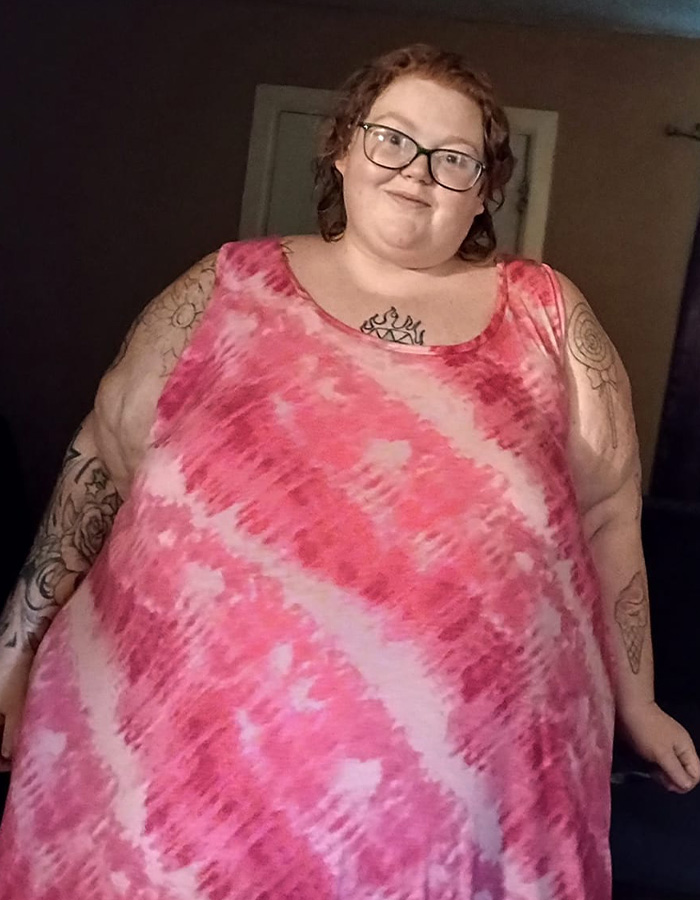Dolly Martinez from My 600-Lb Life wearing glasses and a pink tie-dye dress, smiling indoors with visible tattoos.