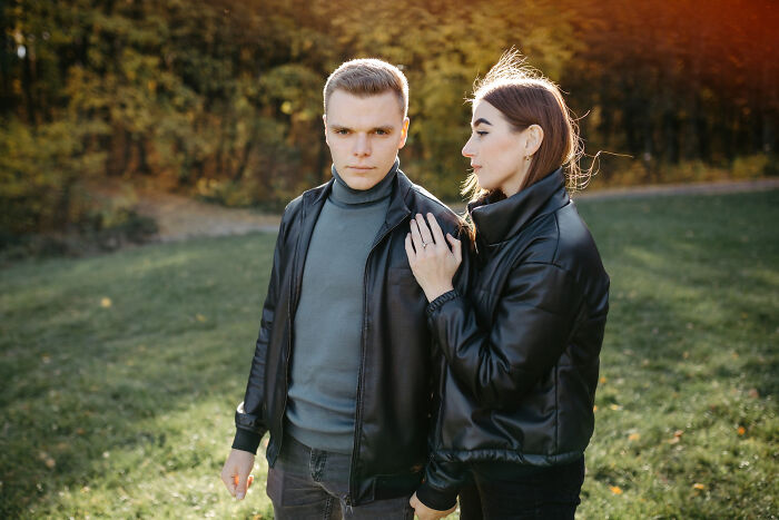 A couple in black leather jackets, standing in a sunny park. He looks at the camera, she gazes at him, depicting how narcissists leave a mark.