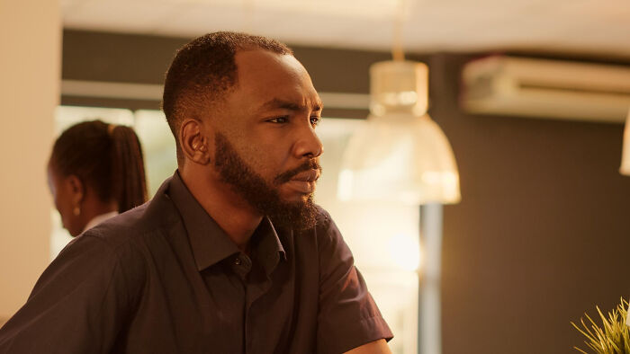 A pensive Black man with a beard, in a dimly lit room. He looks left, leaving a mark on those he meets.