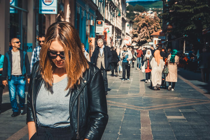 A woman with sunglasses and a leather jacket walks on a busy street, looking down, possibly reflecting on diagnosed narcissists.