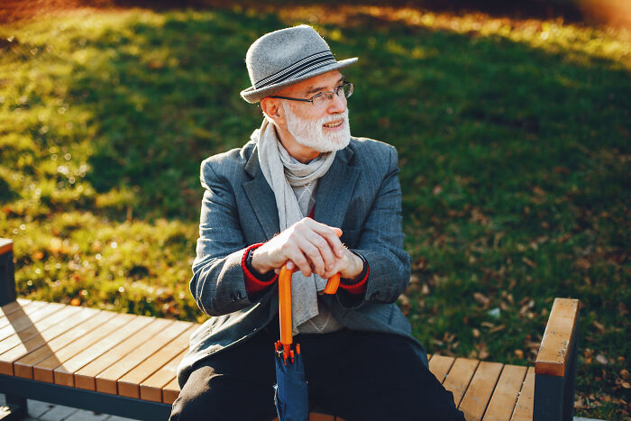 Elderly man in a hat and scarf sits on a bench, holding an umbrella. He looks right, contemplating narcissists.