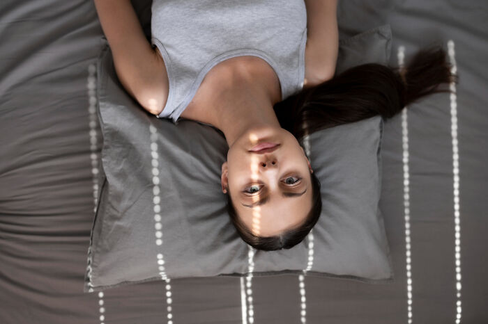 A young woman lying on a gray bed, head on a pillow, looking at the camera. Light stripes hit her face, suggesting she's affected by narcissists.