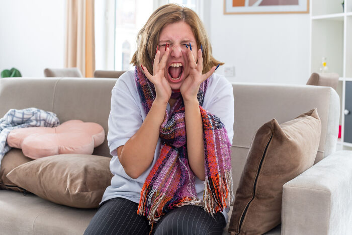 A young woman, wearing a colorful scarf, sits on a couch, screaming with hands near her face, expressing the mark of narcissists.